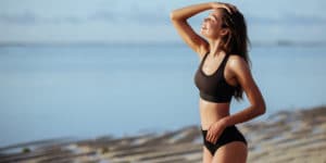 Outdoor shot of smiling young female model in bikini standing against blue sky. Woman having fun out on a summer day.